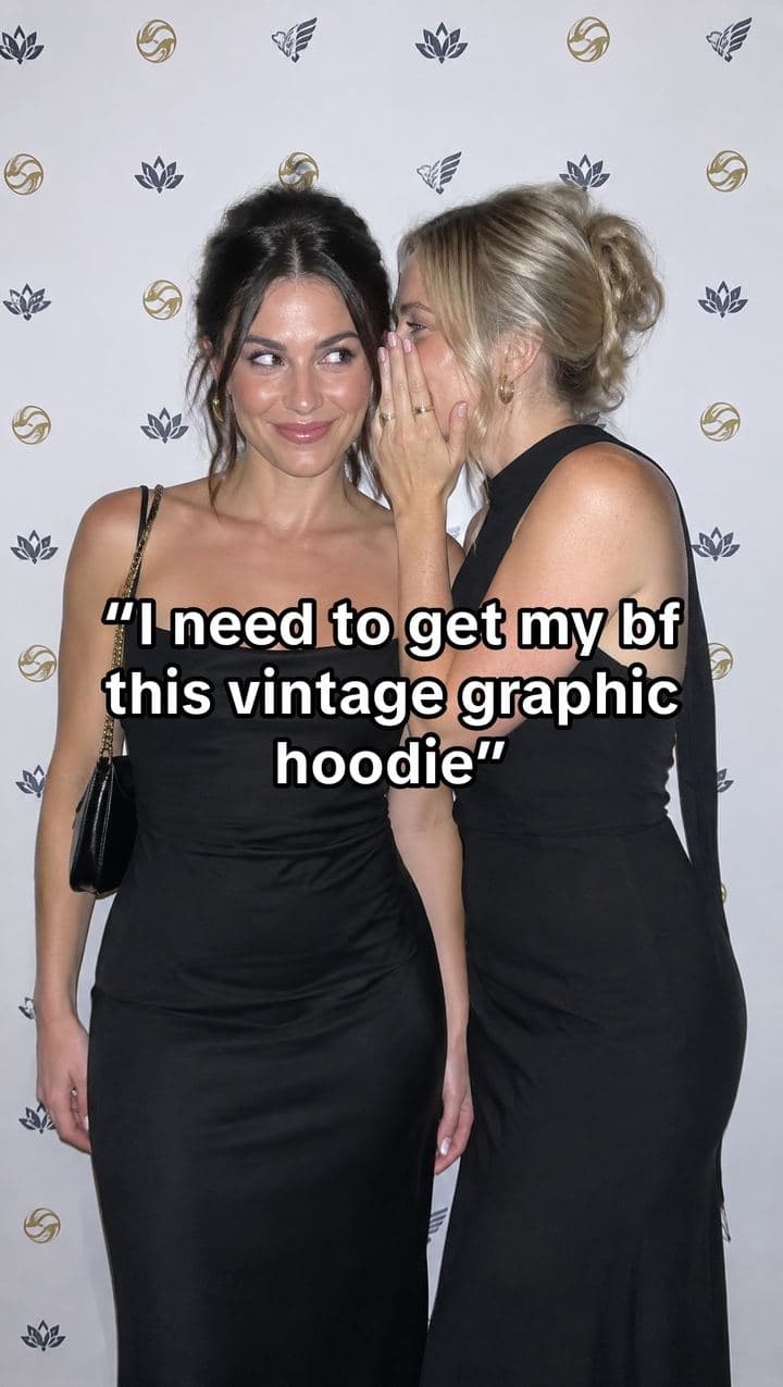 Two friends whispering at a press wall with the text: I need to get my bf this vintage graphic hoodie
